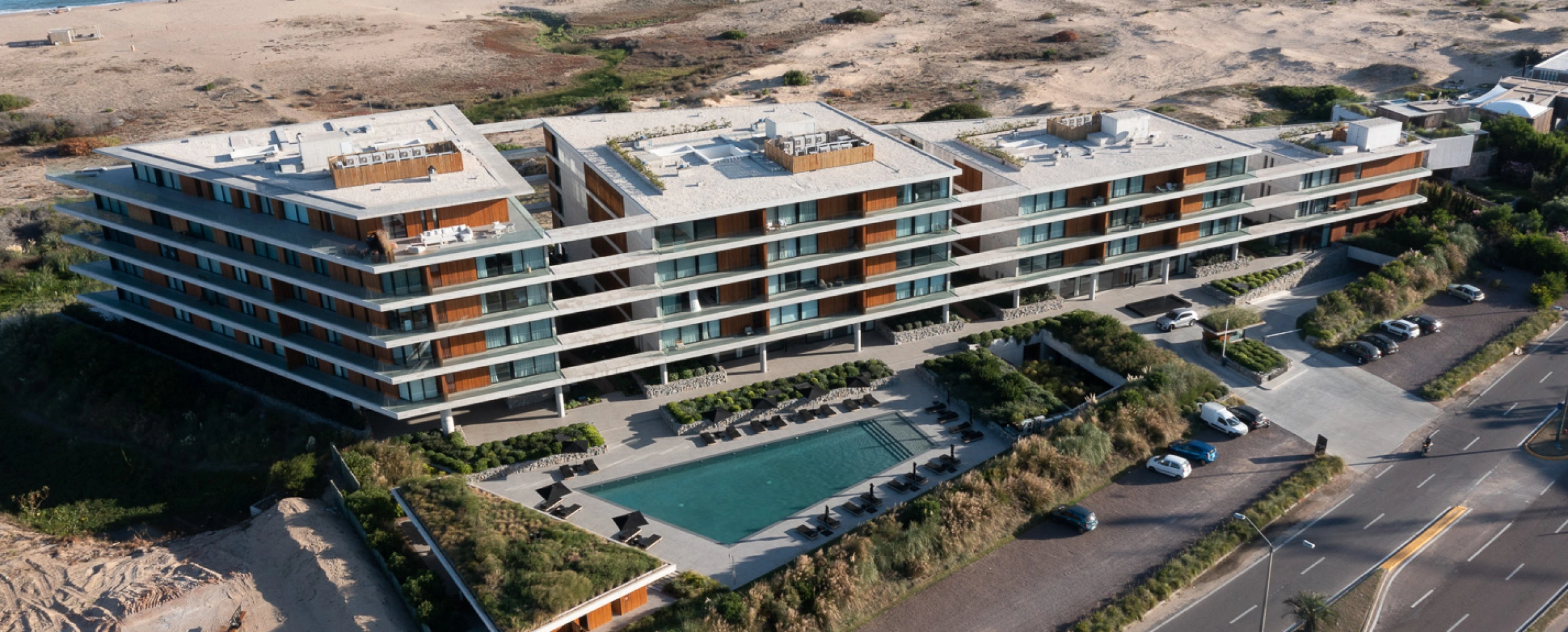 The Colette Residences: Lujo y confort a orillas del mar Vista aérea de un complejo residencial de lujo en la playa. El edificio, de diseño moderno, cuenta con múltiples pisos, una piscina, áreas verdes y estacionamiento. Se encuentra ubicado en una zona costera, rodeado de dunas y vegetación.