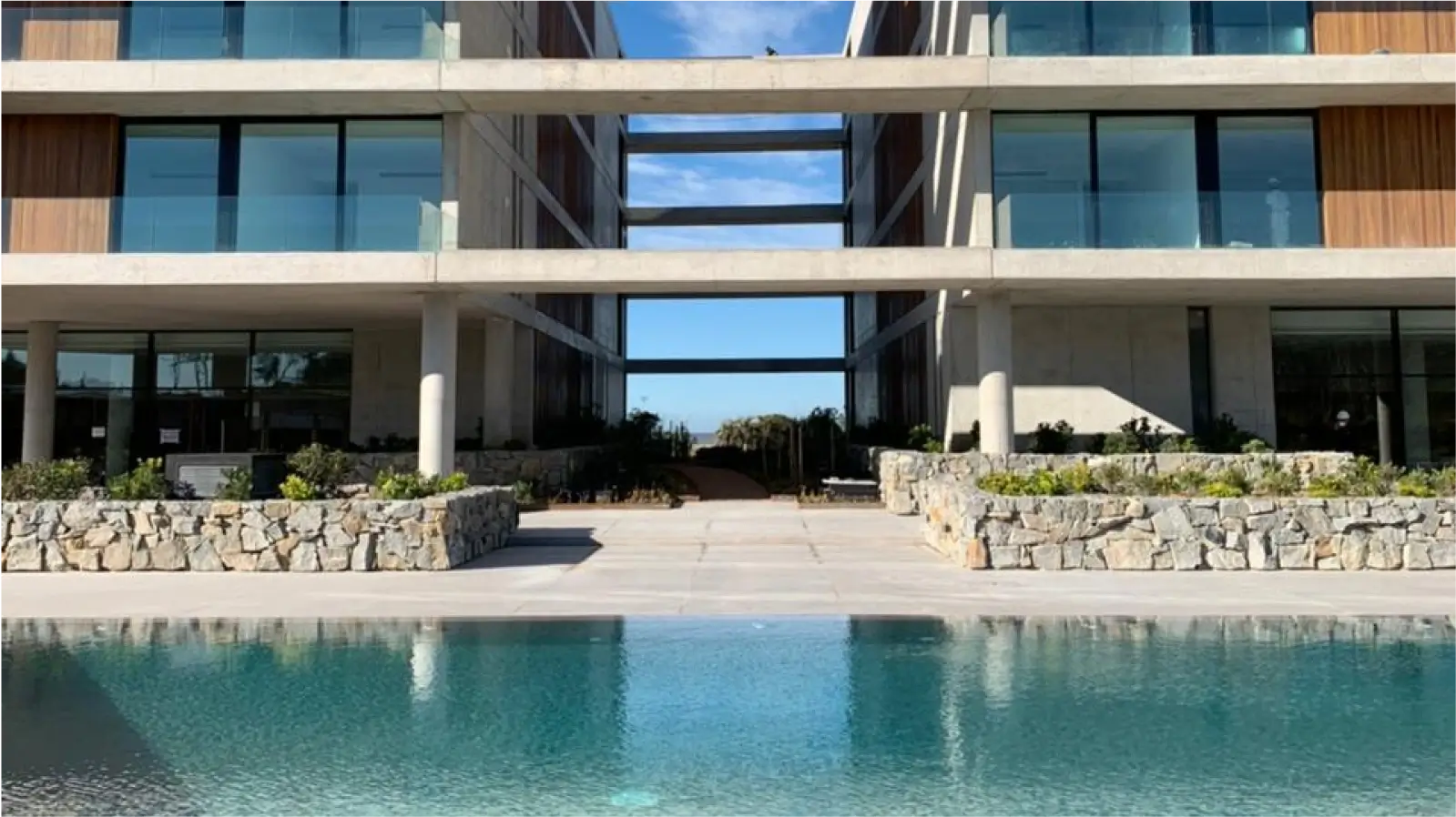 Complejo de apartamentos modernos con piscina y vista al mar. El edificio cuenta con grandes ventanales, balcones y un diseño minimalista. La piscina está rodeada de vegetación y se encuentra en un patio interior.