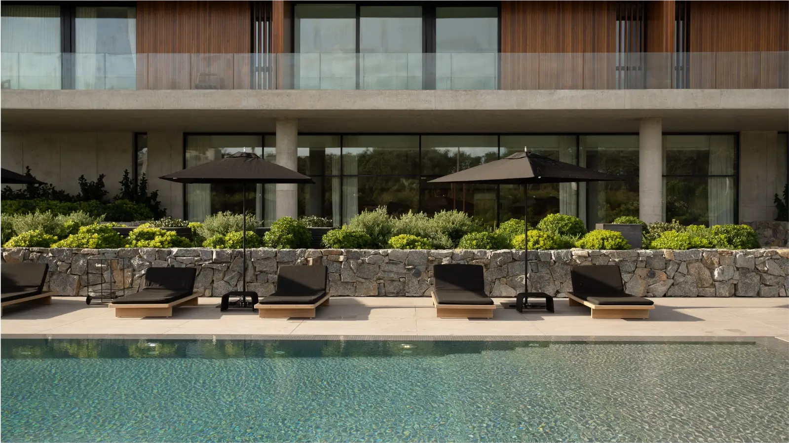 Piscina al aire libre en un complejo hotelero moderno. La piscina está rodeada de tumbonas y sombrillas, y cuenta con un muro de piedra natural como fondo. El edificio principal del hotel, con grandes ventanales y balcones, se encuentra al fondo.