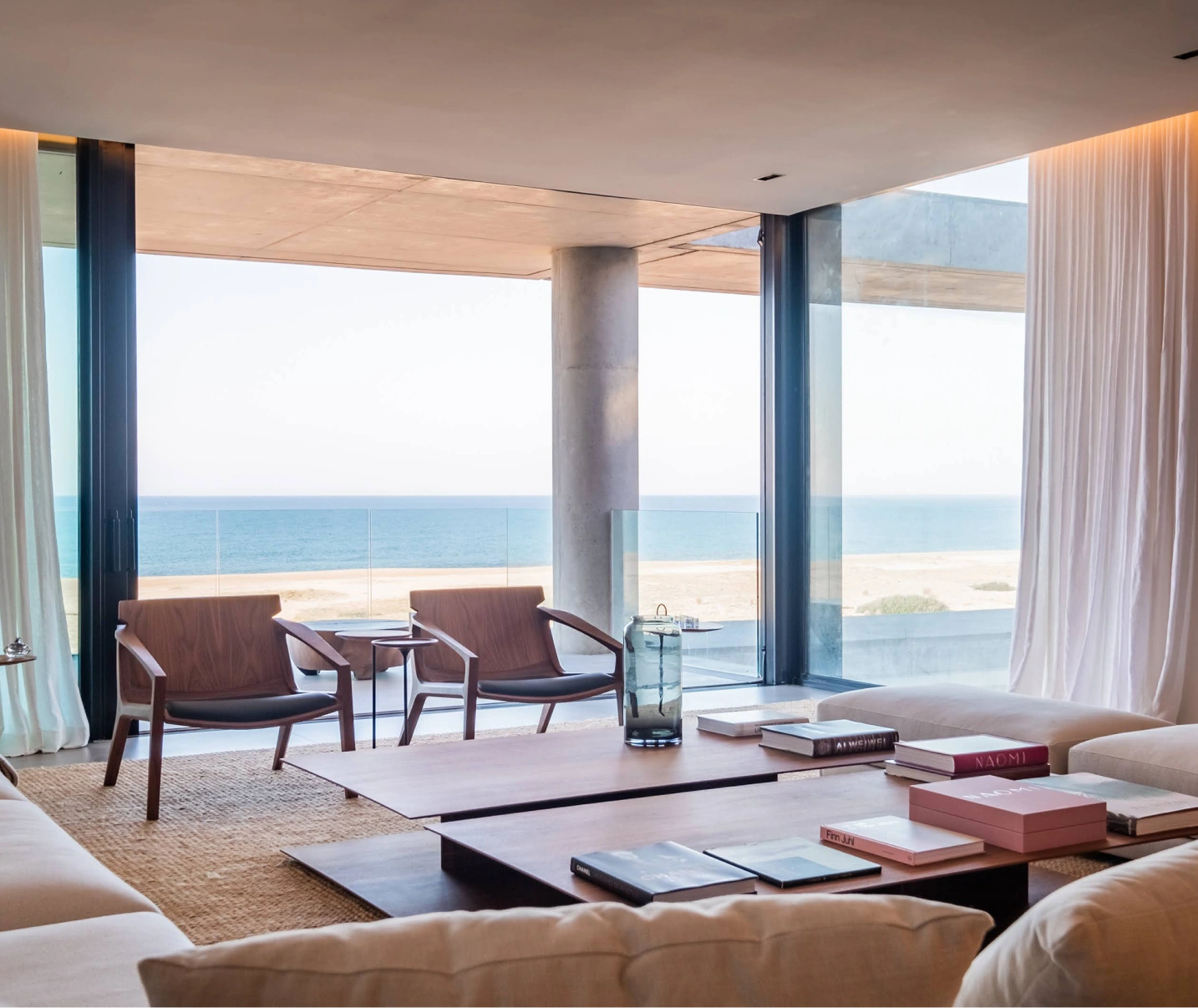 El lujo de vivir frente al mar: Salón con vistas panorámicas Amplio salón con vistas panorámicas al mar. El salón cuenta con grandes ventanales que permiten el ingreso de luz natural, un sofá y sillones tapizados en tonos neutros, una mesa de centro de madera y una alfombra de fibras naturales. El diseño es moderno y minimalista.